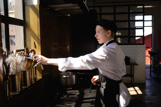 Side view of a Caucasian female chef reaching for a utensil standing in a pot sitting on a window sill in the sun in a restaurant kitchen.