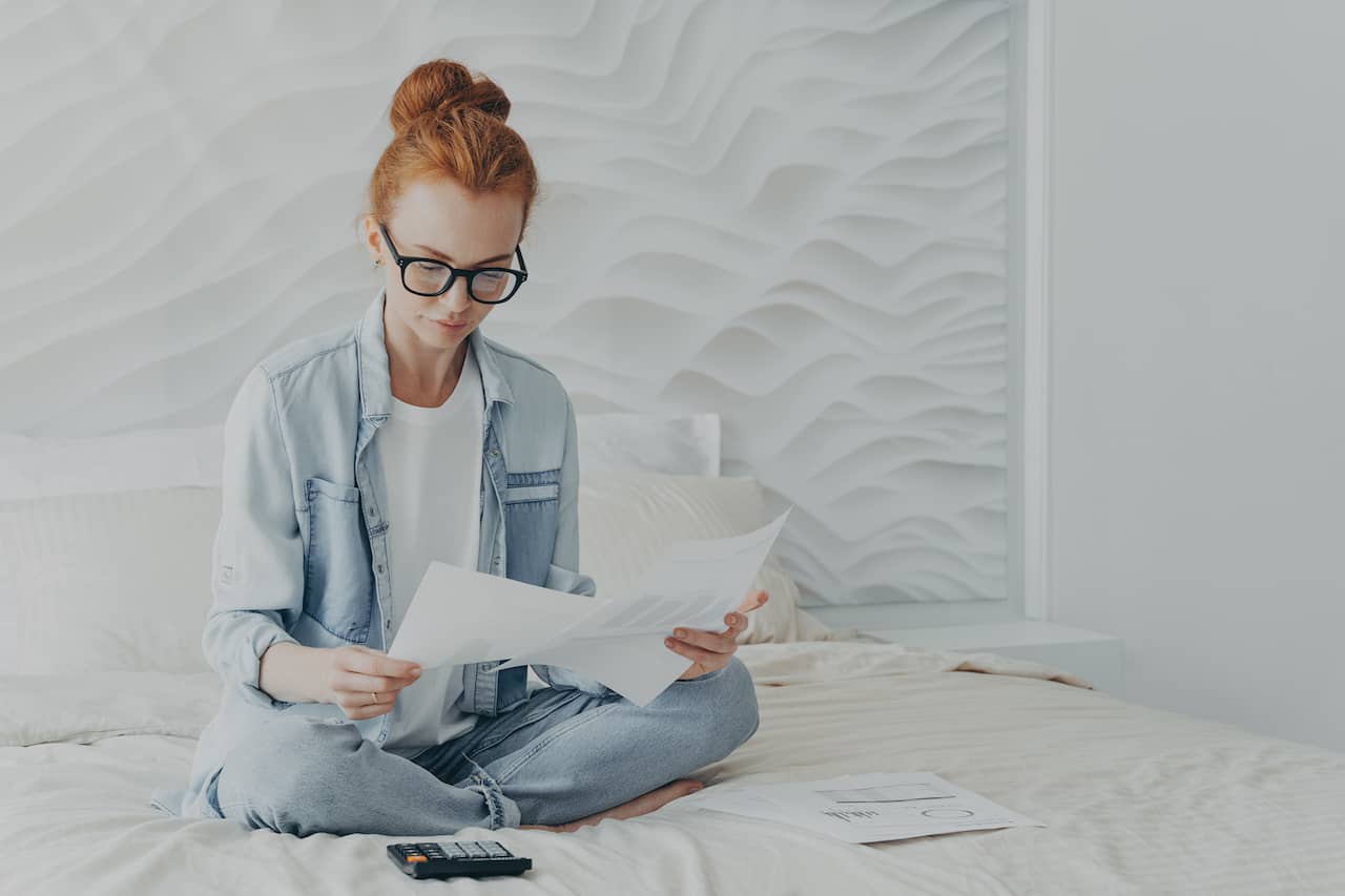 Employee sitting on bed looking at payroll documents.